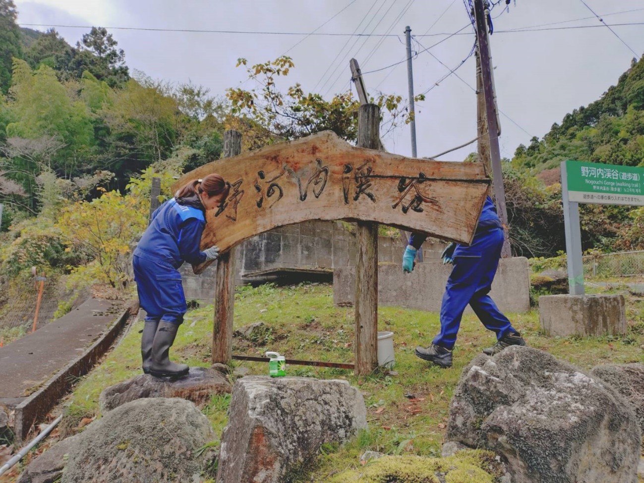 渓谷入り口の書道の達人が書いた案内板を磨く早良区役所職員 苔で見えなくなっていた 左手は三瀬峠越えの国道263号線